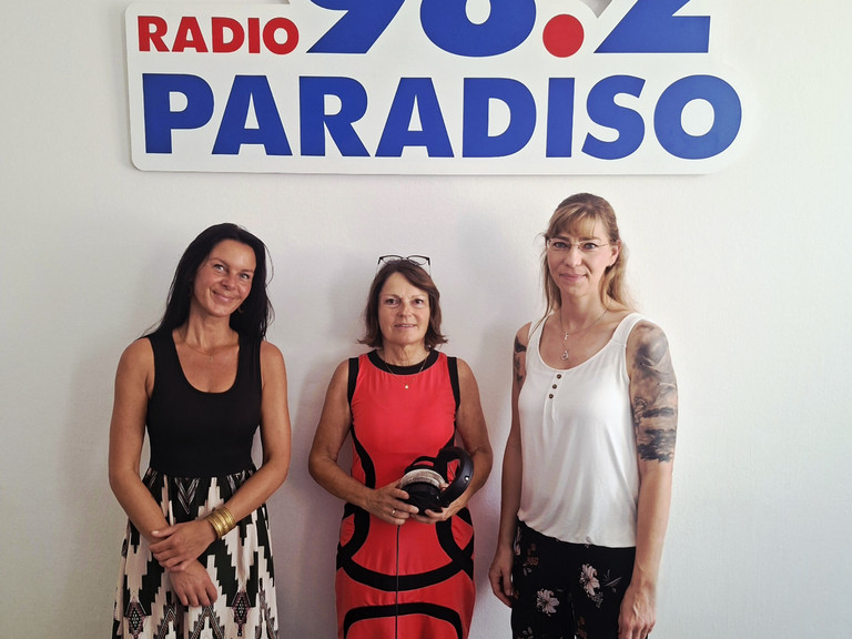 Kunsttherapeutin Sabrina Müller, Radiomoderatorin Julia Nogli und Pflegedienstleitung Stefanie Micklitza stehen unter dem Logo von Radio Paradiso bei der Sendung über die Ermöglichung wertvoller und erfüllter Momente am Lebensende im Diakonie Hospiz Woltersdorf.