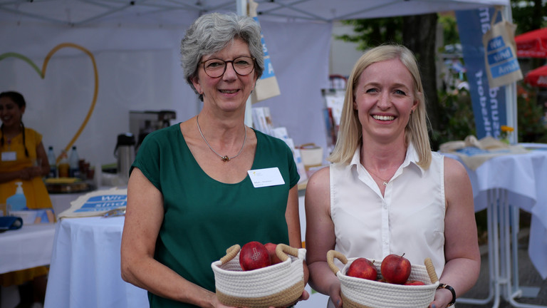 Zwei Mitarbeiterinnen des Amalie Sieveking Krankenhauses begrüßen Gäste mit Äpfeln am Stand des Gesundheitscampus Volksdorf beim Volksdorfer Stadtteilfest in Hamburg-Volksdorf