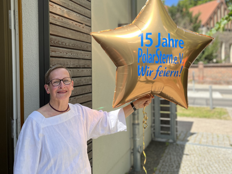 Cornelia Heine hält vor blauem Himmel einen goldfarbenen, sternförmigen Luftballon mit der blauen Aufschrift "15 Jahre PolarStern e.V. - Wir feiern!" in die Kamera bei der Feier zum 15-jährigen Bestehen des Fördervereins „Polarstern“ Neuenhagen e. V. zur Unterstützung des Diakonie Hospiz Woltersdorf im Gemeindesaal der Ev. Verheißungskirchengemeinde Neuenhagen-Dahlwitz.