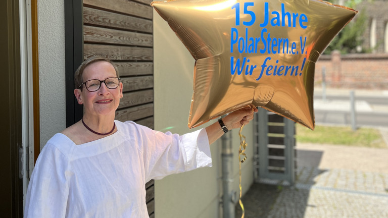 Cornelia Heine hält vor blauem Himmel einen goldfarbenen, sternförmigen Luftballon mit der blauen Aufschrift "15 Jahre PolarStern e.V. - Wir feiern!" in die Kamera bei der Feier zum 15-jährigen Bestehen des Fördervereins „Polarstern“ Neuenhagen e. V. zur Unterstützung des Diakonie Hospiz Woltersdorf im Gemeindesaal der Ev. Verheißungskirchengemeinde Neuenhagen-Dahlwitz.