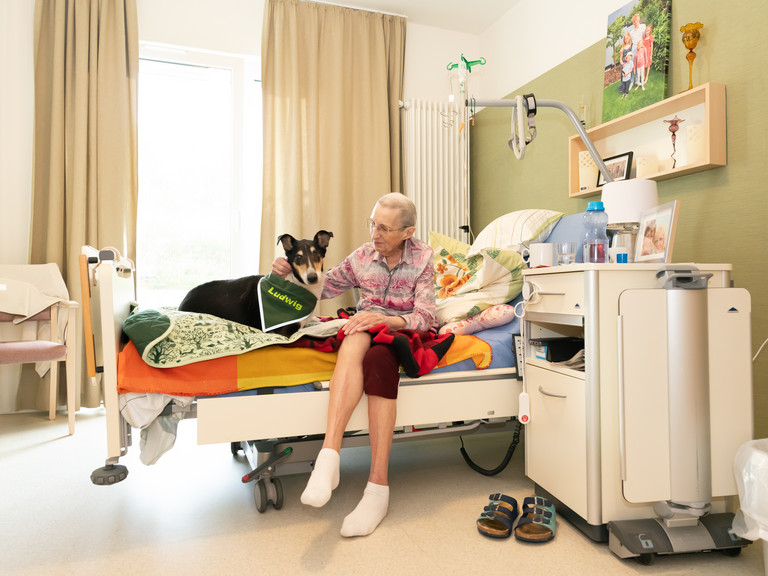 Ein Hospizgast sitzt auf der Bettkante und streichelt einen Therapiehund im Diakonie Hospiz Wolters